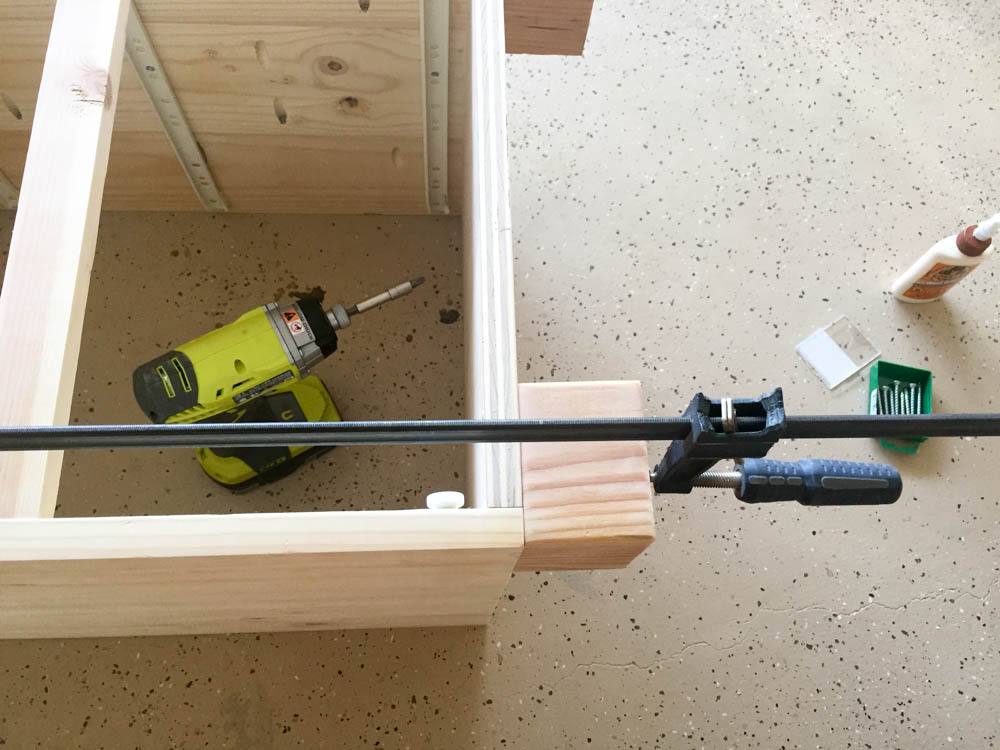 Bar clamp holding a 4x4 wooden leg against the corner of the nightstand base while being secured with construction screws
