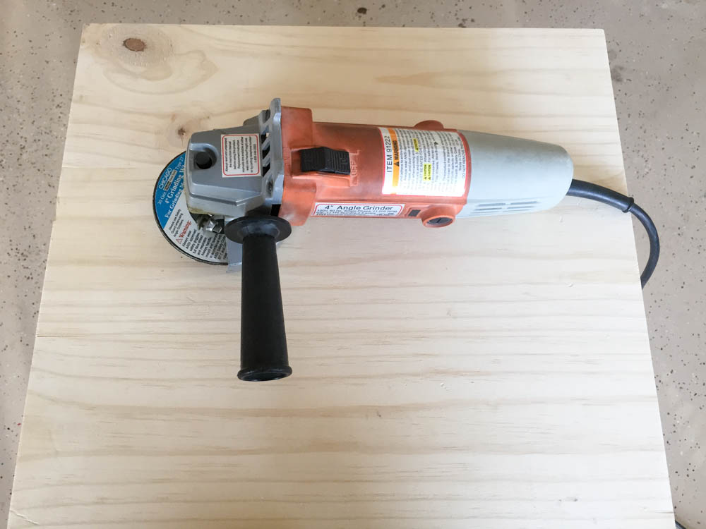 Orange and gray 4-inch angle grinder with grinding disc resting on unfinished pine board before distressing technique