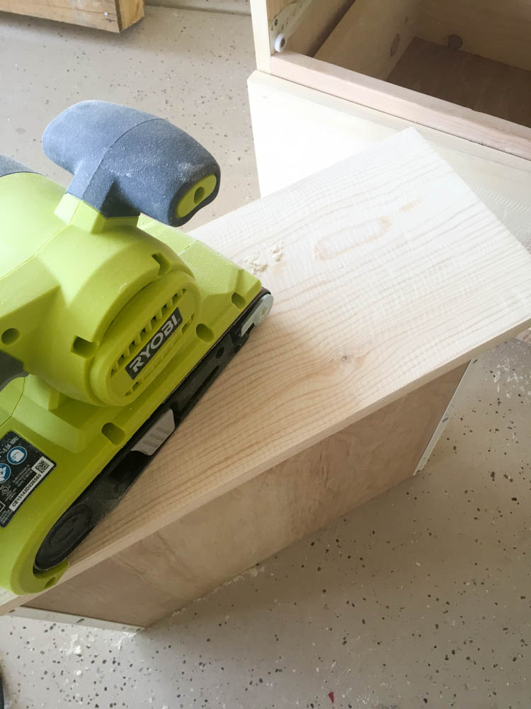 Ryobi belt sander running along the top edge of unfinished pine nightstand to smooth faux saw-mark distressing