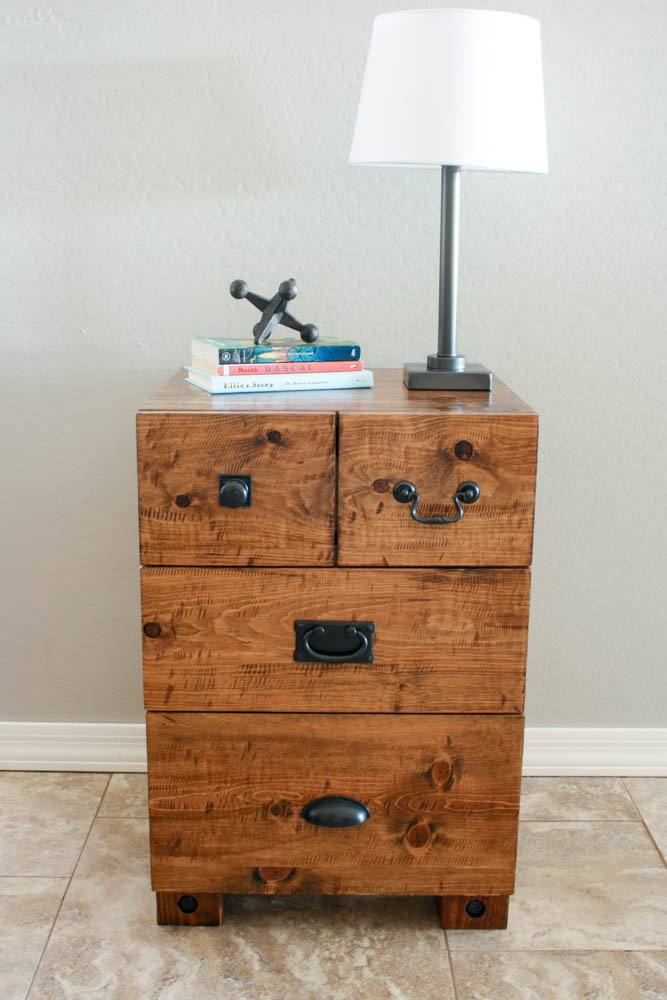 Straight-on view of completed DIY rustic nightstand with three drawers, decorative lag bolt legs, and mixed black hardware styled with lamp and books against a neutral wall