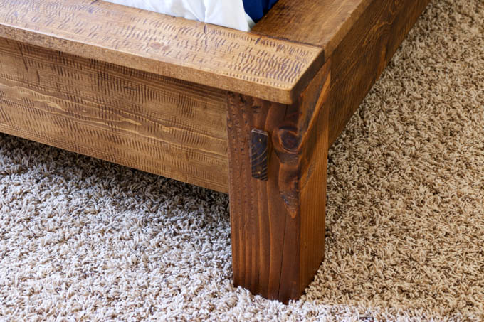 Close-up of distressed wood footboard corner showing mortise-and-tenon strip detail and 4x4 leg on a DIY rustic bed frame