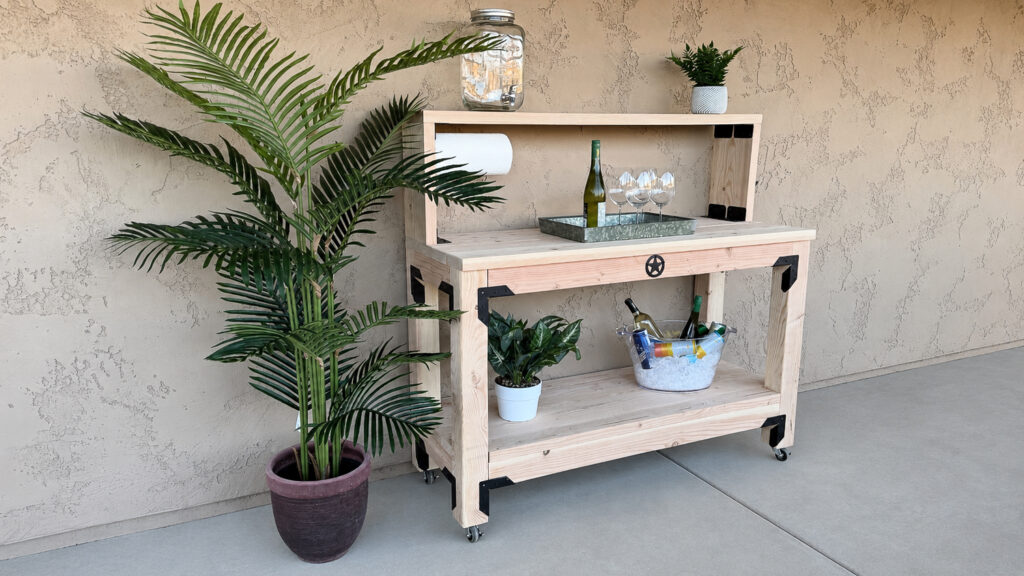 Angled view of the DIY outdoor console table showing the locking casters, black metal brackets, and lower shelf styled with a plant and drink bucket