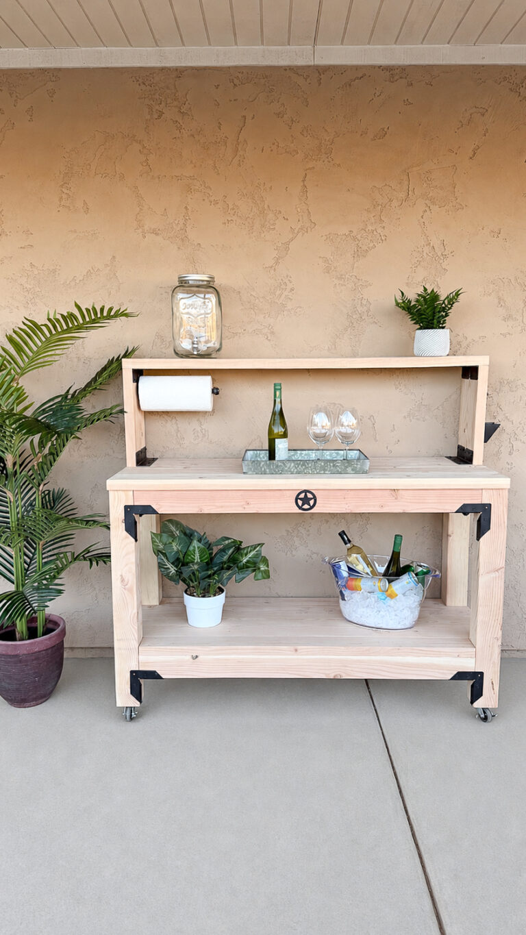 Full-length front view of the finished outdoor console table against a stucco wall, set up as a drink station with a beverage dispenser, wine, and glasses