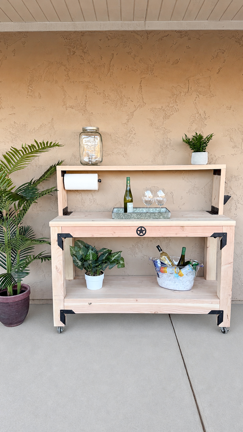 Full-length front view of the finished outdoor console table against a stucco wall, set up as a drink station with a beverage dispenser, wine, and glasses