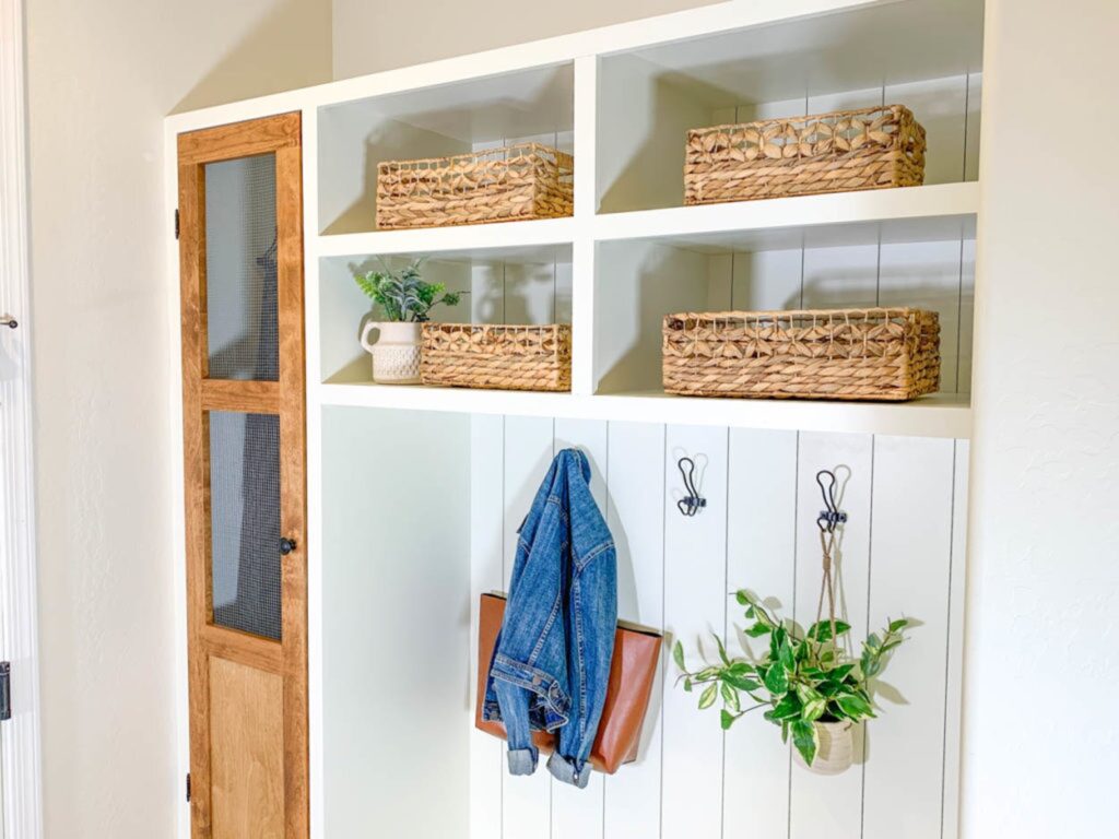 Upper portion of a custom mudroom built-in with open storage cubbies, woven baskets, hooks and a wood cabinet door