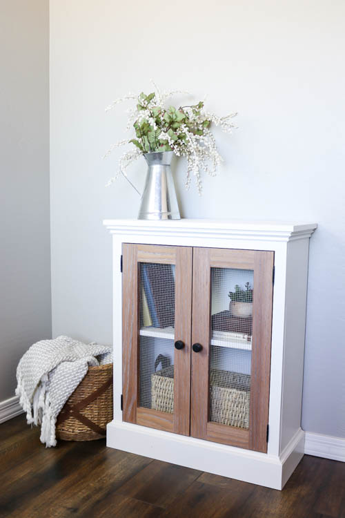 Finished two-tone cabinet with white painted body and weathered oak mesh doors styled in a living room corner