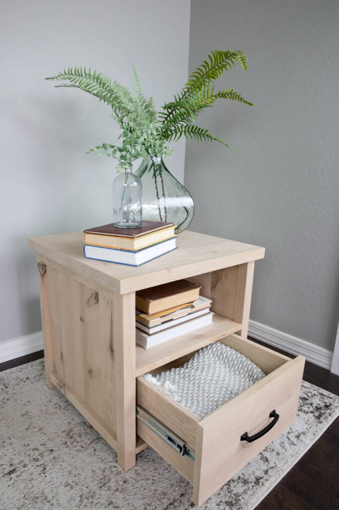 Natural wood DIY side table with the storage drawer open, holding a folded knit blanket, next to an open book shelf